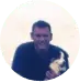  A smiling man holding a small dog, framed in a circular border against a pale background.