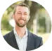  A man with a beard, smiling outdoors, with a blurred green background suggesting a natural, leafy setting.